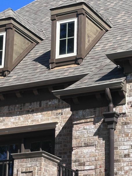 Gable Trim Replacement After/Before Close-up of a home’s roof with dormer windows and brick exterior.