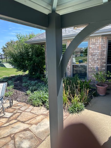 Gazebo View of a patio with plants and a brick house, under a shaded structure.